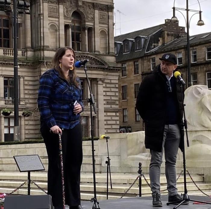 Emma Roddick MSP and another speaker standing on a stage in George Square, Glasgow. Emma is holding a walking stick and speaking into a microphone with a blue cover. The other speaker wears a cap and uses a microphone with a yellow cover. Buildings and part of the square’s architecture are visible in the background.