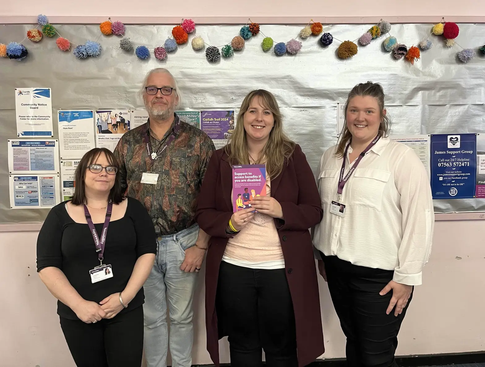 Emma Roddick MSP stands with three members of the local VoiceAbility team in front of a bulletin board, holding a booklet about support for accessing Social Security Scotland benefits.