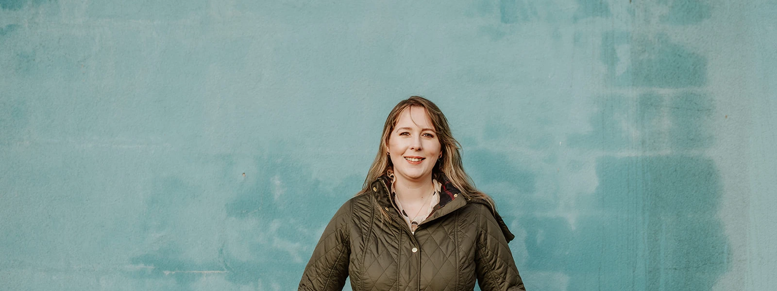 Emma Roddick MSP standing against a textured blue wall, wearing a dark green quilted jacket