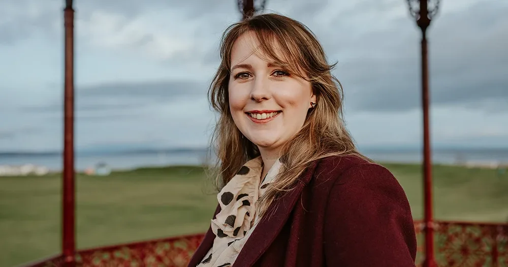 A picture of Emma Roddick MSP smiling, wearing a maroon coat and patterned scarf, she stands in a red gazebo with grass and cloudy sky in the background.