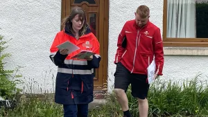 Emma Roddick MSP standing with a postal worker outside a house. Both are wearing postal uniforms and holding letters.