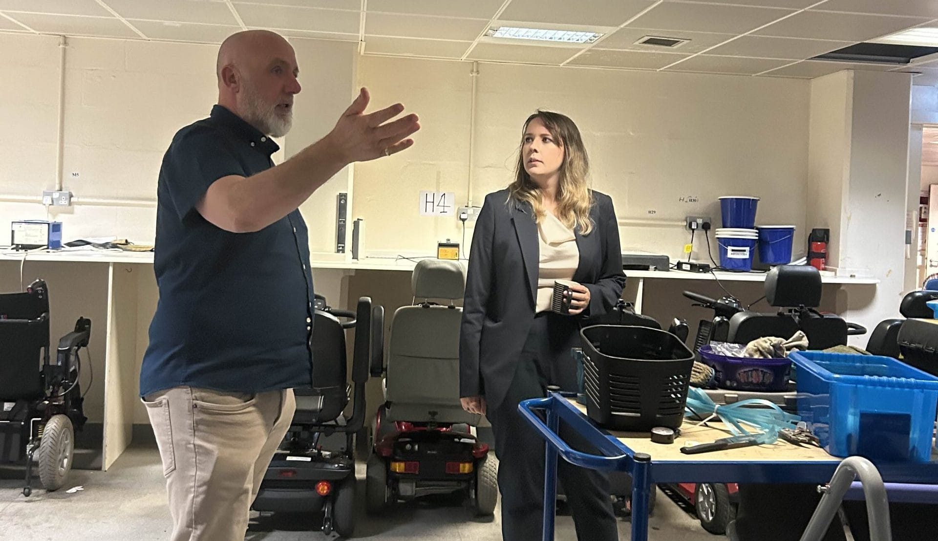 Emma Roddick MSP in a workshop surrounded by rows of mobility scooters; one gestures while speaking, the other listens holding a mug beside a trolley of tools and baskets