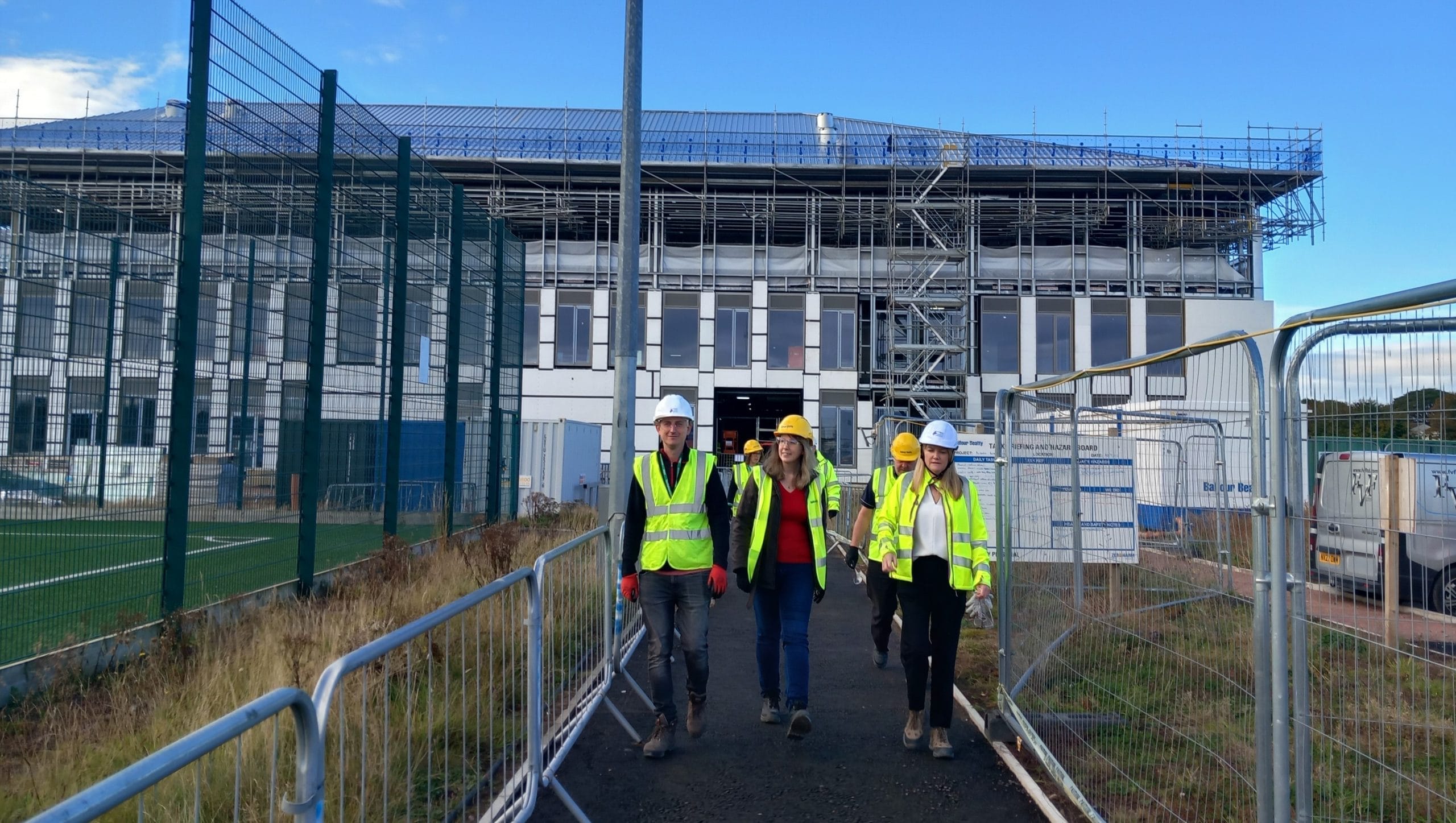 Emma with high vis vest, joined by contractors, outside school building that is under construction
