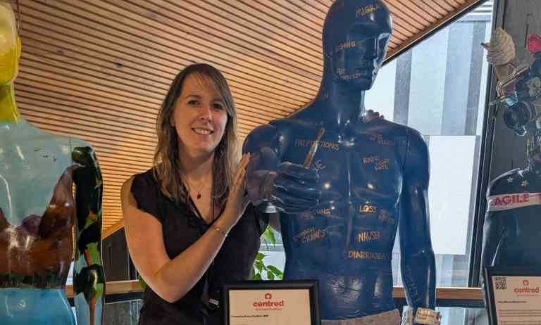 Emma stands in Eden Court, posing with a mannequin, painted blue and wearing grey joggers. He is painted with physical symptoms of mental illness in gold lettering, and holds a paintbrush.