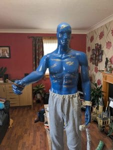 Body of Work stands in a living room, with more of him visible, including a crutch in his left hand which is plastered with mental health medication packaging.