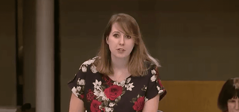 Emma speaks in parliament, wearing a black top with a red and white floral pattern.