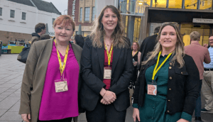 Emma Harper, Emma Roddick, and Karen Adam stand outside SNP conference, all wearing yellow lanyards and suits