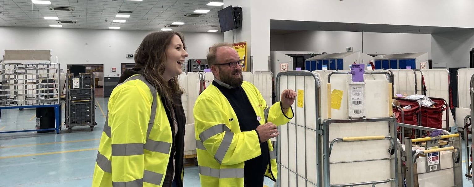 Emma, in a hi-vis jacket, tours a sorting office with a man who is pointing off-camera and speaking to her