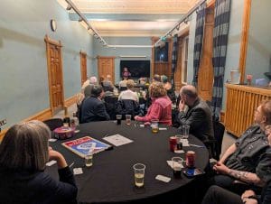 A crowd of SNP members, friends, and family sit at tables, watching Fiona who speaks at the far end of the room.