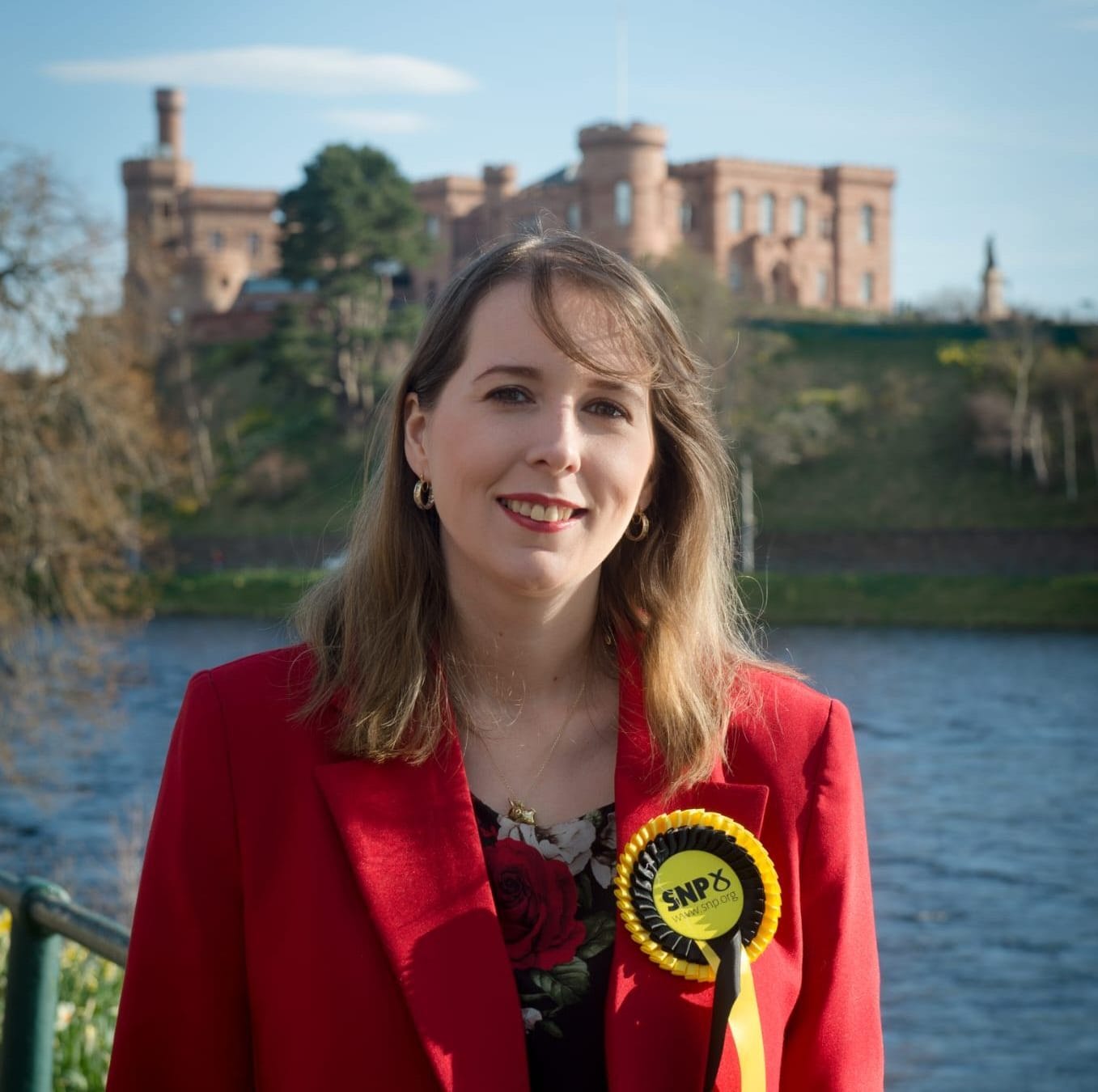 Emma wears a red blazer and SNP rosette, the river and Inverness Castle behind her.